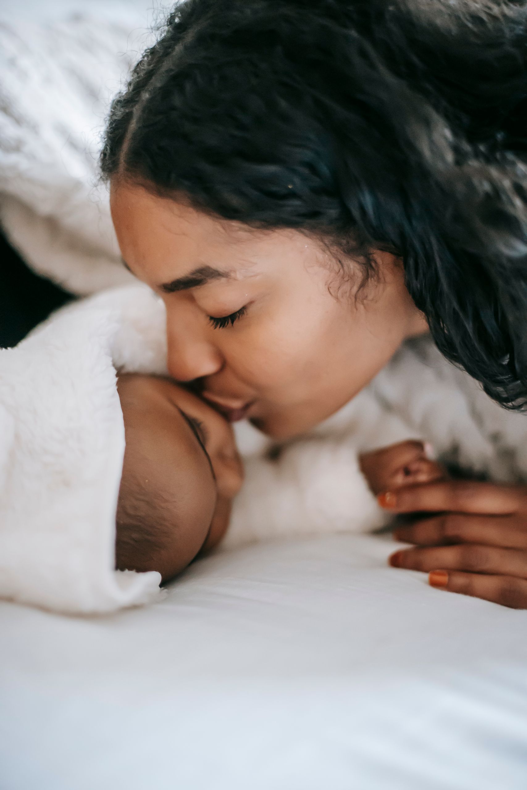 Calm ethnic mother kissing little baby lying on bed and sleeping at home in daylight