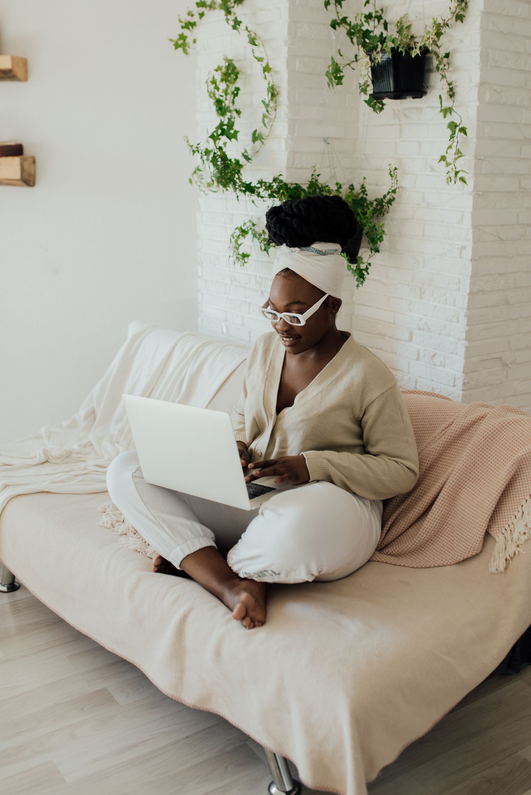 African American woman sitting on a sofa using a laptop, wearing eyeglasses and a headwrap in a bright living room.